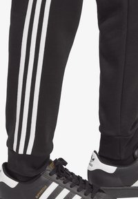 Svarta sweatpants med tre vita ränder längs sidorna, kombinerade med svarta och vita Adidas Superstar-sneakers.