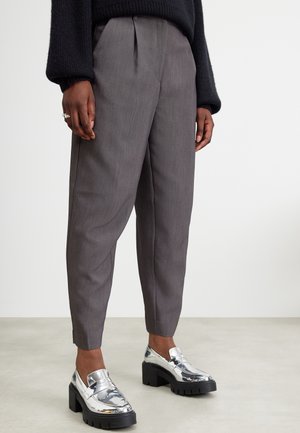 Person, der bærer grå skræddersyede bukser, sort langærmet sweater og skinnende sølv chunky hælede loafers på lys gulvbaggrund.