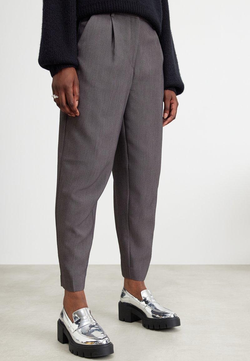 Personne portant un pantalon gris sur mesure, un pull noir à manches longues et des mocassins argentés brillants à talons épais sur un fond de sol clair.