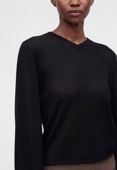 Femme portant un pull noir à manches longues col en V avec une texture tricotée et un pantalon marron sur un fond gris uni.