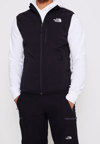 Mann in schwarzem Reißverschluss-Weste und Hose, mit einem weißen Langarm-Hoodie darunter, alles mit dem Logo von The North Face versehen.