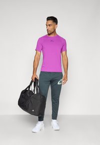 T-shirt de sport violet à manches courtes, logo noir, associé à un pantalon vert foncé avec un logo vert clair. Sac de sport noir, chaussures blanches.