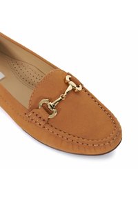 Tan suède moccasin met een gouden hardware accent op de wreef, voorzien van ton-sur-ton stiksels en een ronde neusdesign. Het interieur is licht geperforeerd.