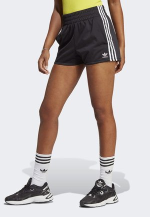 Benen in zwarte Adidas-short met witte strepen, witte Adidas sokken met zwarte strepen en zwarte Adidas sneakers op een effen achtergrond.