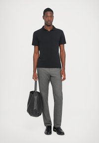Mann steht da, trägt ein schwarzes Poloshirt und graue Hosen und hält eine schwarze Ledertasche in seiner rechten Hand.