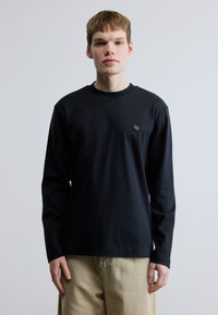 Črna majica z dolgimi rokavi iz bombaža, z okroglim izrezom in majhno logotipno značko na levi prsni strani. Nošena z bež hlačami.