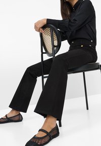 Mustat leveälahkeiset housut yhdistettynä mustaan napitettavaan paitaan ja mustiin verkkomateriaalisiin ballerinoihin. Tuolissa on pyöreä rottinkipintainen istuin ja tyylikäs musta runko.