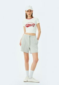 Camiseta corta blanca con detalles rojos y gráfico de "California", combinada con pantalones cortos gris claro. El modelo lleva calcetines y zapatillas blancas.