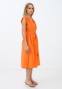 Oranje katoenen jurk met een V-hals, mouwloos ontwerp, een taille met riem en een geplooide rok. Model draagt bruine sandalen met bandjes.