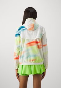 White hooded jacket with multicolored watercolor patterns in blue, green, and orange, featuring an elastic hem. Worn with a bright green skirt.