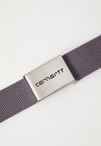 Carhartt öv textúrált sötétlila szőtt szíjjal és ezüstszínű négyzet alakú fémbuckle-val, amelyen gravírozott logó található.