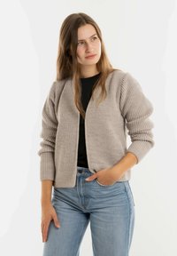 Lichtbruine gebreide cardigan met geribbelde textuur, open voorkant en lange, licht poffende mouwen. Gepaard met een donkere top en blauwe jeans.