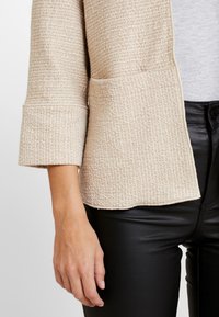 Beige texturovaná bunda s jemným tkaným vzorem, tříčtvrteční rukávy a volný střih, zkombinovaná s černými úzkými kalhotami.