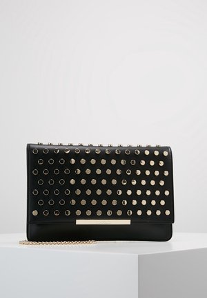 Pochette rectangulaire noire avec des clous ronds dorés et une bandoulière chaîne dorée posée sur une surface blanche sur un fond uni.
