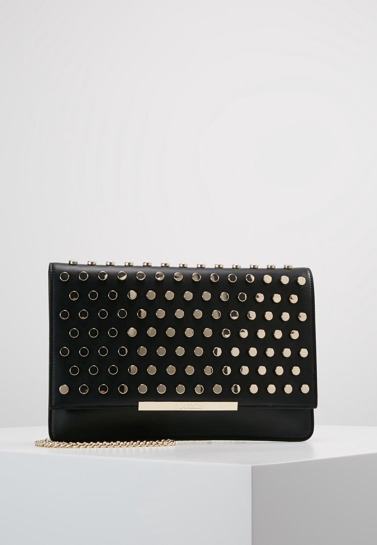 Pochette rectangulaire noire avec des clous ronds dorés et une bandoulière chaîne dorée posée sur une surface blanche sur un fond uni.