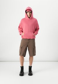 Haut à capuche rose avec une poche avant, associé à un short cargo marron avec des poches latérales. Des bottes noires complètent la tenue.