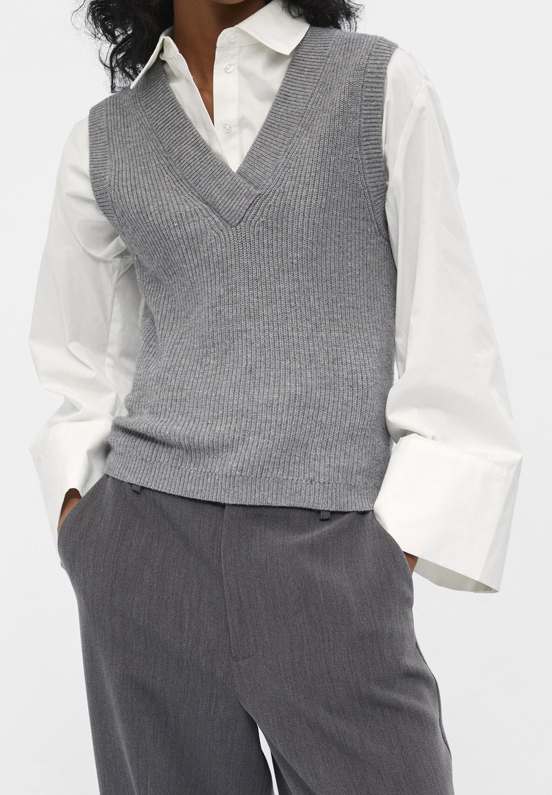 Veste grise en maille avec un col en V, portée sur une chemise blanche à col, accompagnée d'un pantalon gris texturé.