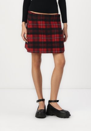 Femme portant une jupe trapèze à carreaux rouge et noir, un haut noir à manches longues et des chaussures Mary Jane noires à semelle épaisse avec des brides à la cheville.