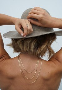 Beige Filzhut rückwärts getragen; drei goldene Ketten hängen über den Rücken, mit einer strukturierten Oberfläche an den Ketten. Nahaufnahme von Händen, die den Hut anpassen.