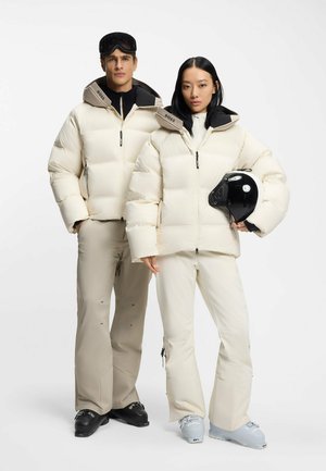 Dwóch modeli w beżowych kurtkach narciarskich i spodniach; jeden trzyma czarny kask, obaj noszą zimowe buty na jednolitym białym tle.