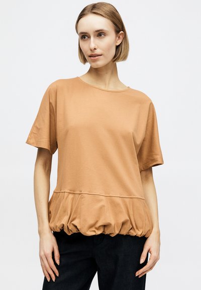 Γυναίκα που φοράει μια χαλαρή κορυφή με κοντά μανίκια σε καμηλό χρώμα με ρυτιδωμένη χαμηλή ραφή, σε συνδυασμό με μαύρο παντελόνι, στέκεται μπροστά σε λευκό φόντο.