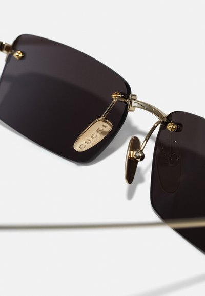 Ochelari de soare negri, de formă rectangulară, cu accesorii metalice aurii, care prezintă un panou cu logo pe podul nasului și tampoane nazale ajustabile.