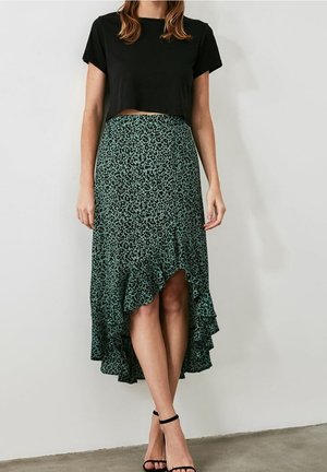 Femme portant un t-shirt court noir, une jupe midi asymétrique imprimé léopard vert et des talons noirs à bride à la cheville, debout contre un mur blanc.