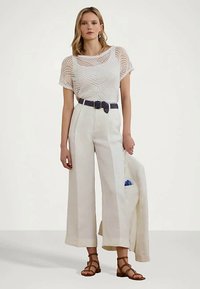 Moteris vilki baltą raštuotą permatomą marškinėlį, aukšto juosmens plačias baltas kelnes su juodu diržu, laiko baltą švarką, rudos sandalais.