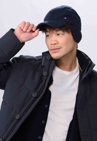 Marineblaue Wollmütze mit Ohrenklappen, weiche Textur, mit Schirm. Getragen mit einer schwarzen Daunenjacke und einem hellen T-Shirt darunter.