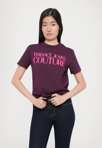 Violetinė medvilnės marškinėliai su ryškiai rožiniu tekstu "VERSACE JEANS COUTURE", derinami su tamsiai mėlynomis džinsomis, pabrėžiančiomis aptemptą siluetą.