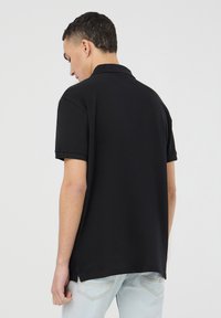 Polo shirt nera con maniche corte, realizzata in tessuto morbido. Presenta un colletto classico ed è caratterizzata da un orlo posteriore leggermente più lungo.