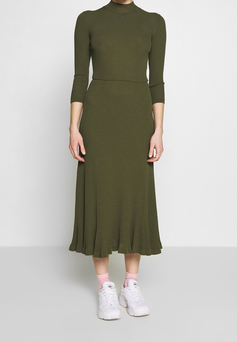 Robe côtelée vert olive avec un col montant, des manches trois-quarts et un ourlet évasé, associée à des baskets blanches et des chaussettes roses.