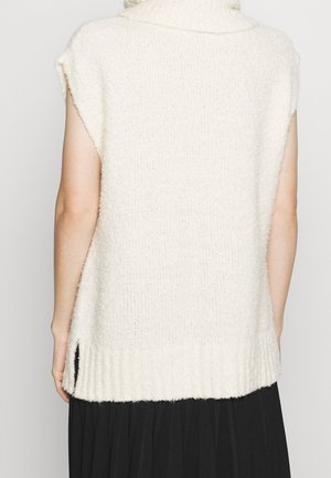 Maglione senza maniche color crema in maglia con collo alto e spacchi laterali, indossato sopra una gonna nera plissettata, visto da dietro.