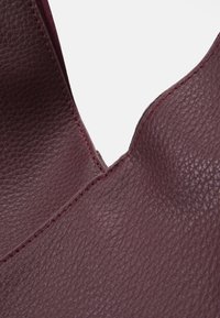 Burgundy nahkakotelo, jossa on teksturoitu pinta, V-muotoinen aukko ja kontrastivärinen punainen ommel reunoilla. Sisäosa on sileä.