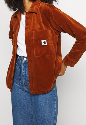 Personne portant une veste en velours côtelé couleur rouille sur une chemise blanche et un jean bleu taille haute, une main sur la hanche.