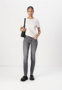 Camiseta gris claro con logotipo, combinada con jeans ajustados grises. El modelo lleva unos mocasines negros gruesos. Un bolso de hombro negro añade contraste.