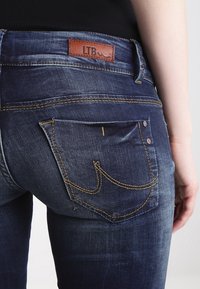 Jeans de mezclilla azul oscuro con una etiqueta de cuero LTB en la cintura. Presenta un patrón de costura único en el bolsillo trasero.
