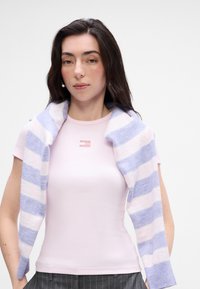 Hele roosa ribidega T-särk väikese logoga rinnus, mille kohal on sinine ja valge triibuline sveter, mis on sõlgade ümber seotud.