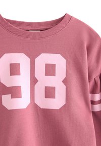 Sweat-shirt rose à col rond, avec un grand imprimé blanc "98" et deux bandes blanches sur les manches. Tissu doux et texturé.