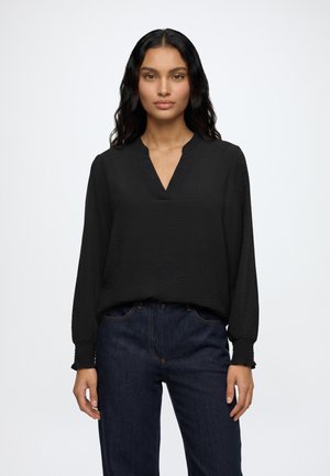 Femme aux longs cheveux noirs portant un chemisier noir à manches longues avec une encolure fendue et un jean bleu foncé, debout devant un fond uni.