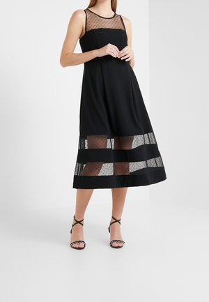 Vrouw draagt een mouwloze zwarte jurk met doorzichtige mesh met polkadots bij de halslijn en zoom, gecombineerd met zwarte open-toe hakken met enkelbandjes.