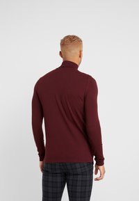 Bordowy, długi rękaw t-shirt z golfem wykonany z miękkiej tkaniny, o dopasowanym kroju i gładkiej fakturze. W zestawieniu z wzorzystymi, w kratę spodniami.