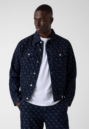 Homme portant une veste en jean bleu foncé à motifs et un pantalon assorti sur un t-shirt blanc, regardant sur le côté avec les mains dans les poches.