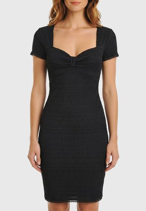 Donna che indossa un vestito nero aderente a maniche corte con scollo quadrato e dettaglio arricciato sul corpetto, in piedi davanti a uno sfondo chiaro e semplice.