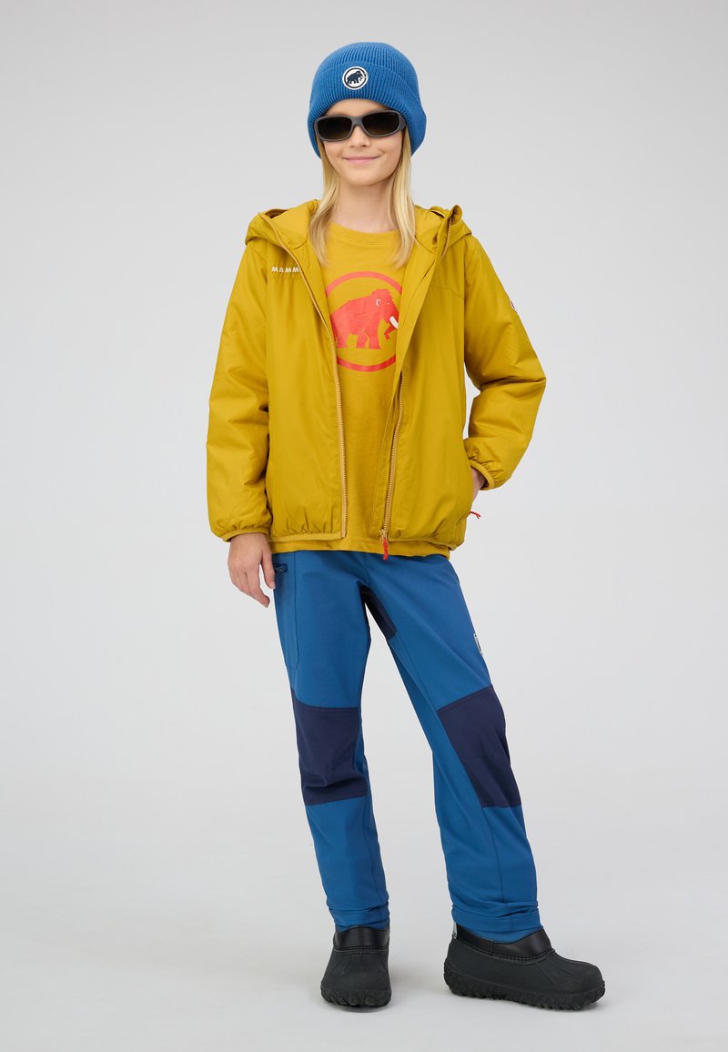Veste zippée jaune, bonnet en tricot bleu, t-shirt graphique avec un éléphant rouge, pantalon bleu avec des accents marine, et chaussures noires.