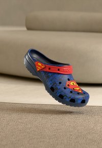 Blue and black clog with red strap featuring yellow and red Superman logos, floating above beige carpet with a sofa in the background.
