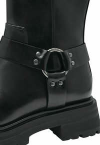 Bota de cuero negra con una correa ancha, hebilla metálica circular y tachuelas decorativas, suela gruesa y texturizada, y perfil elegante.