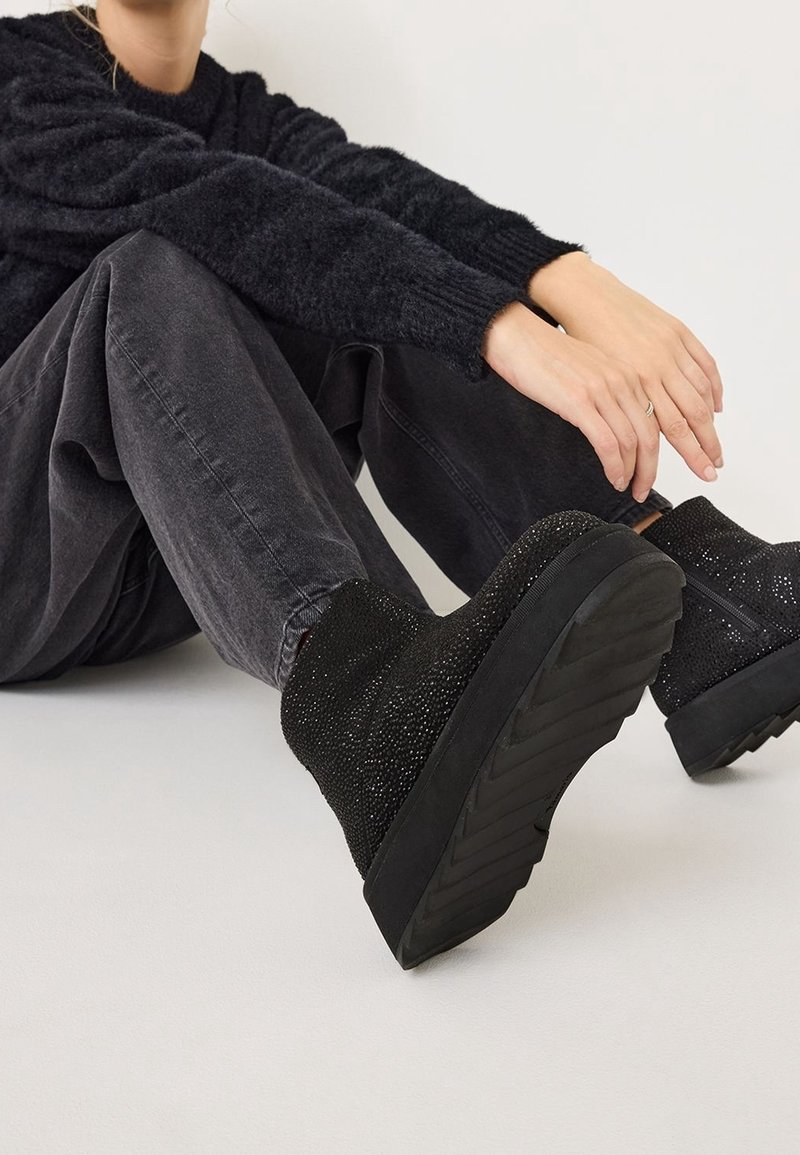 Person in einem schwarzen, flauschigen Pullover, dunklen grauen Jeans und schwarzen, hochhackigen Stiefeletten mit strukturiertem Material, der auf einem hellen Boden sitzt.