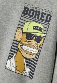Sudadera gris con un mono de dibujos animados sonriendo ampliamente, usando gafas de sol negras y una gorra amarilla, impresa sobre un fondo de rayas.