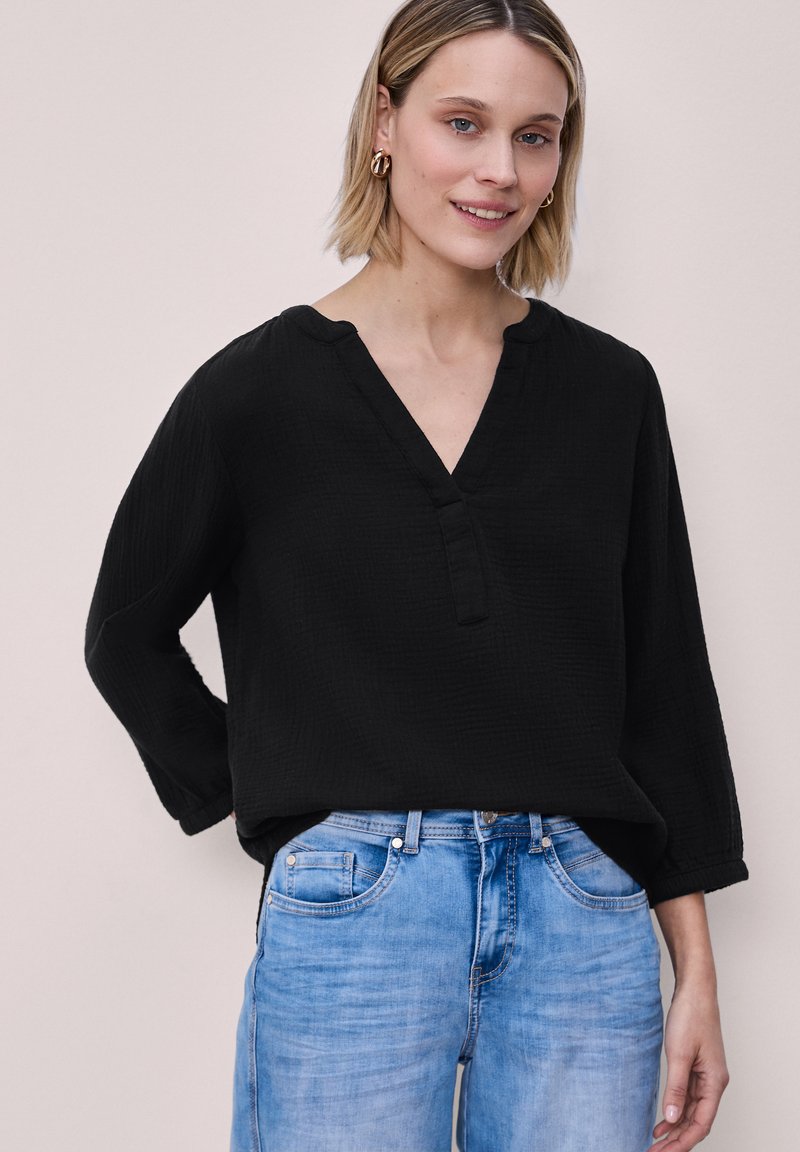 Nainen hymyilee, päällään löysä musta V-kaula-aukollinen paita, jossa on kolmevartiset hihat, ja vaaleansiniset farkut, seisoo yksiväristä taustaa vasten.